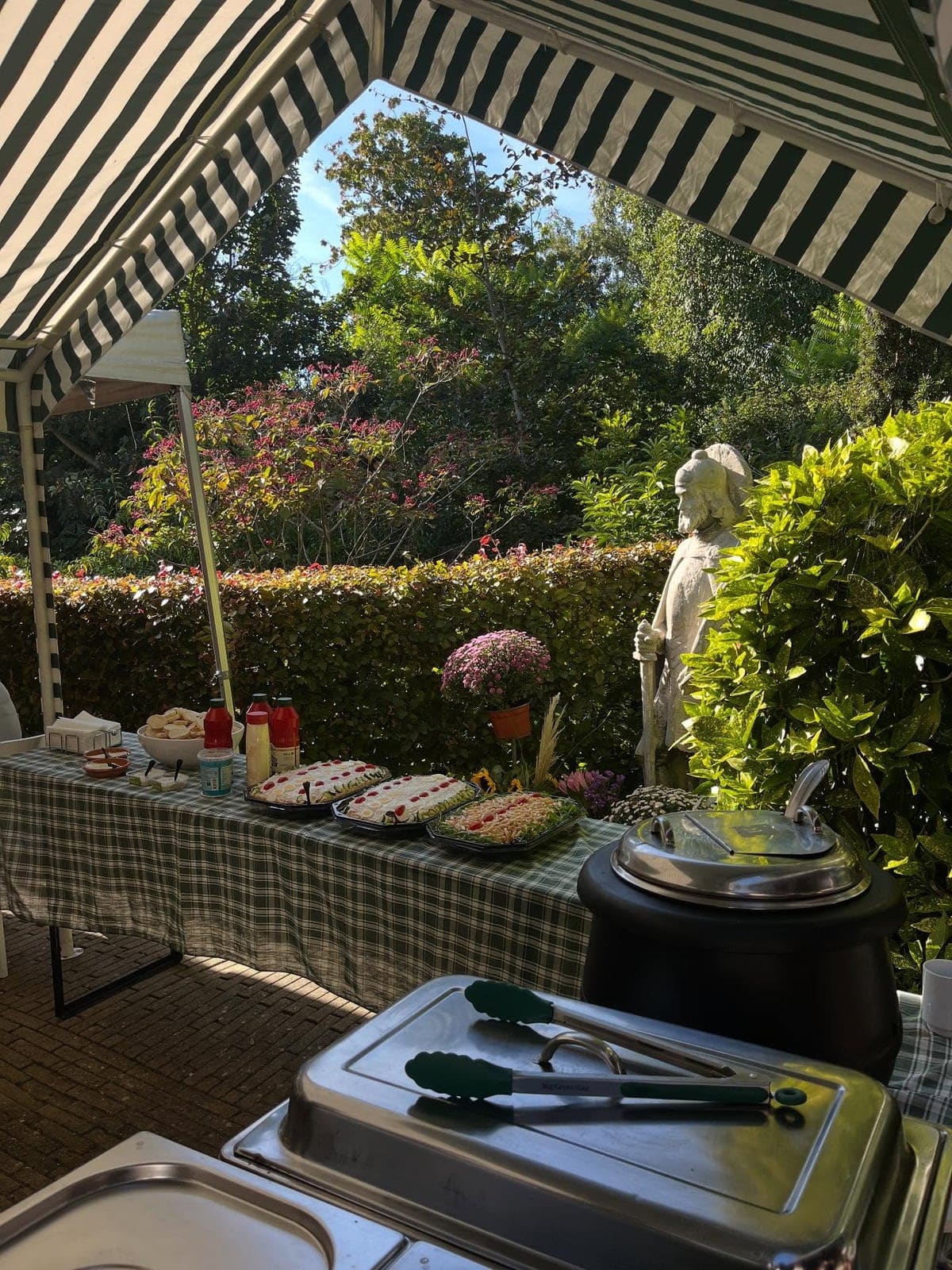 Buffettafel met salades en warmhoudbakken, opgesteld in een tuin onder een gestreepte luifel