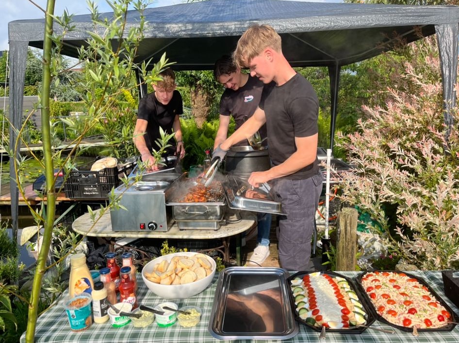 Drie medewerkers van Vuur en Vlam aan de grill onder een partytent in een tuin, met salades en broodjes op tafel